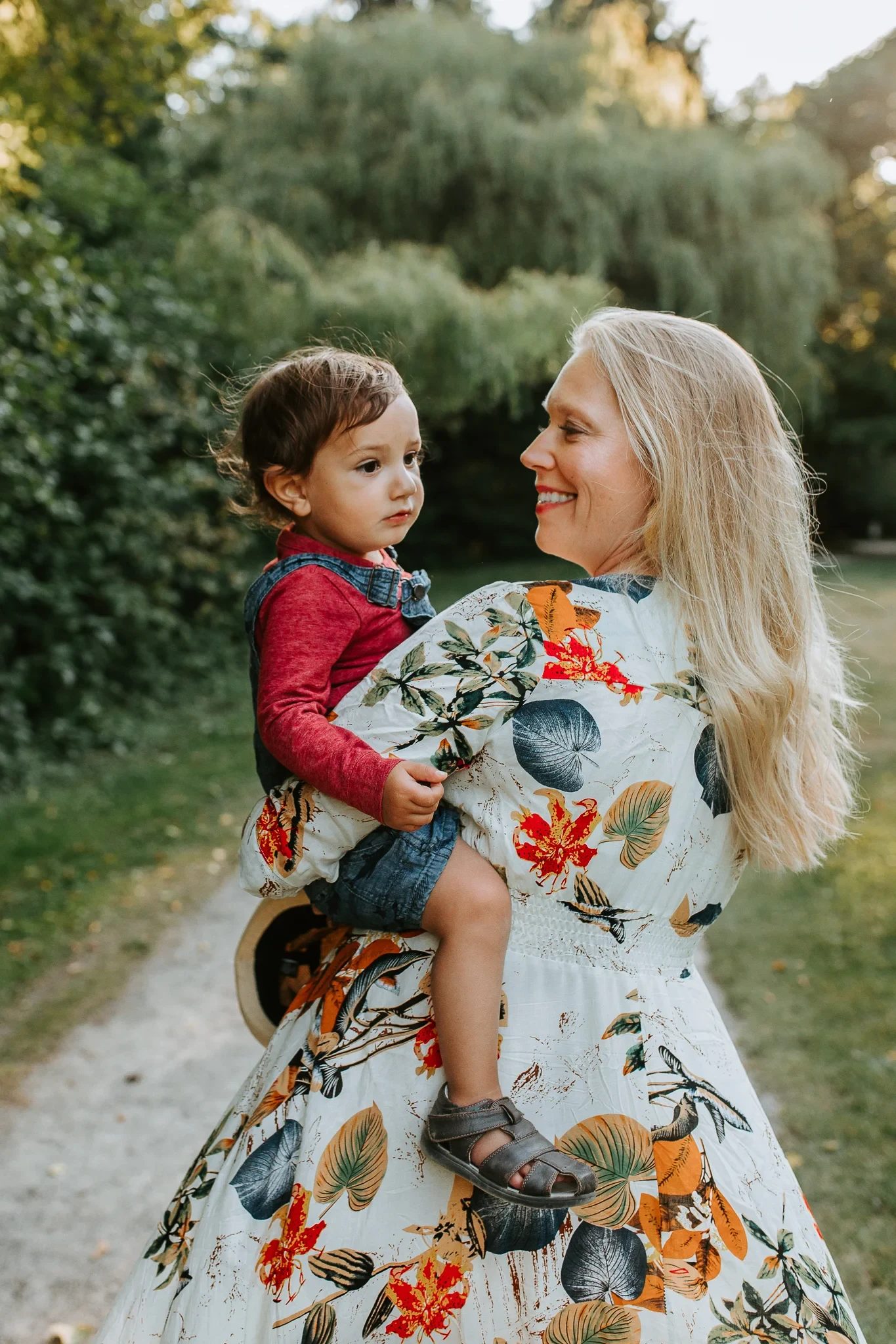 Woman in a floral dress carries a child beside green trees