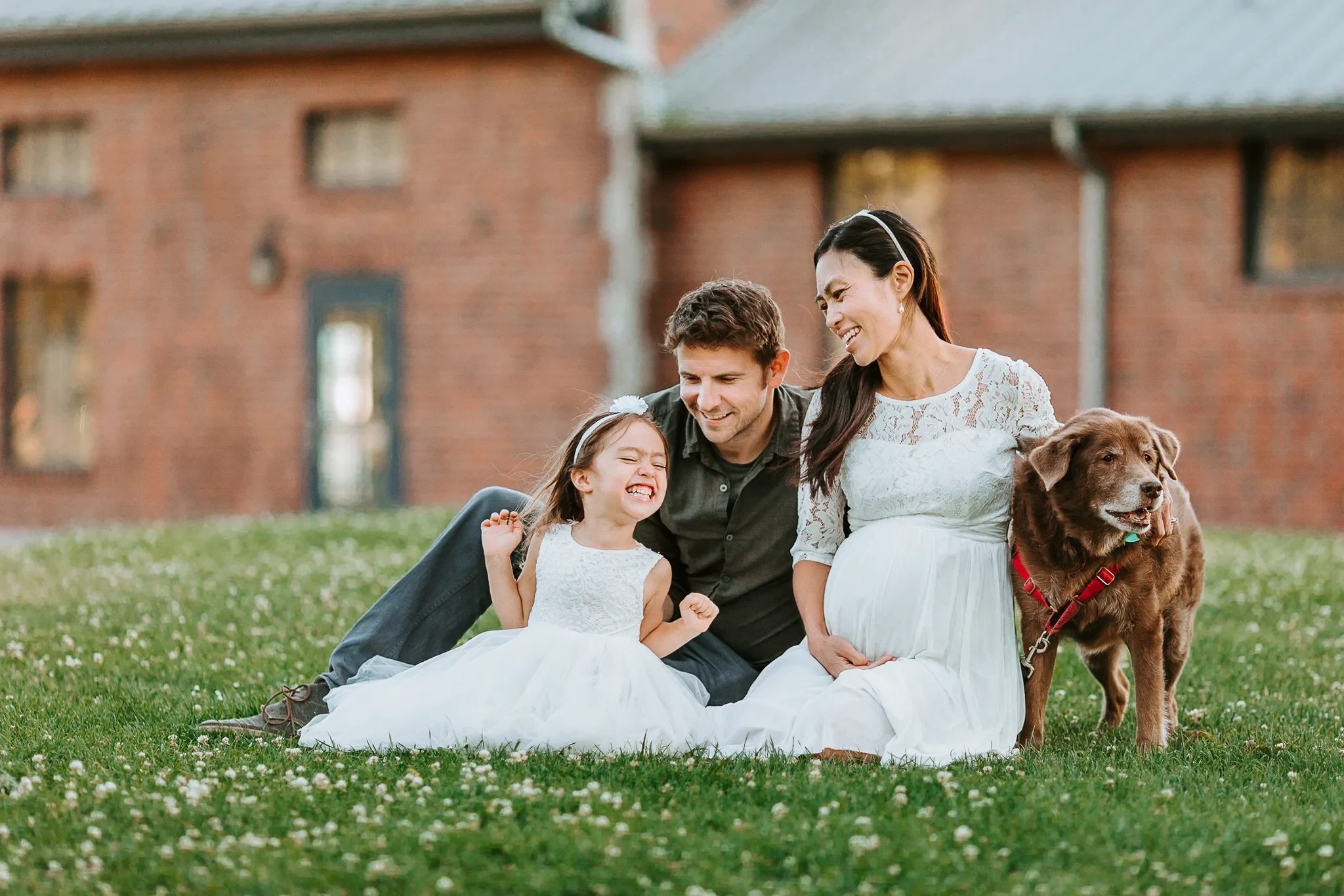 Two adults and a child sit on a lawn beside a dog and a red building