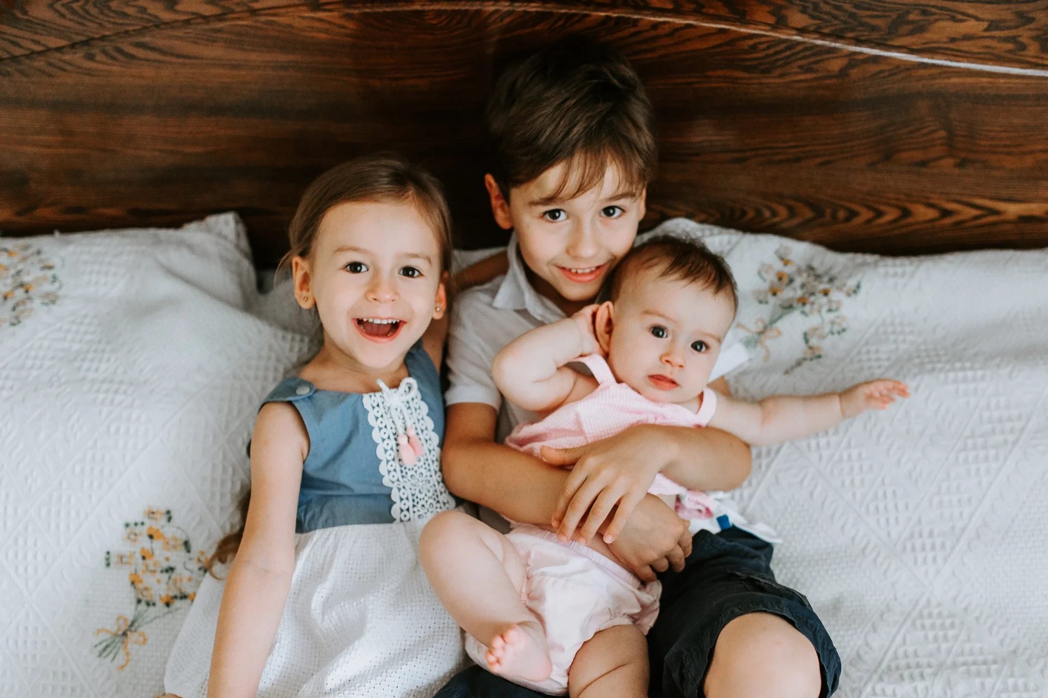 Three children sit close together on a bed with a wooden headboard