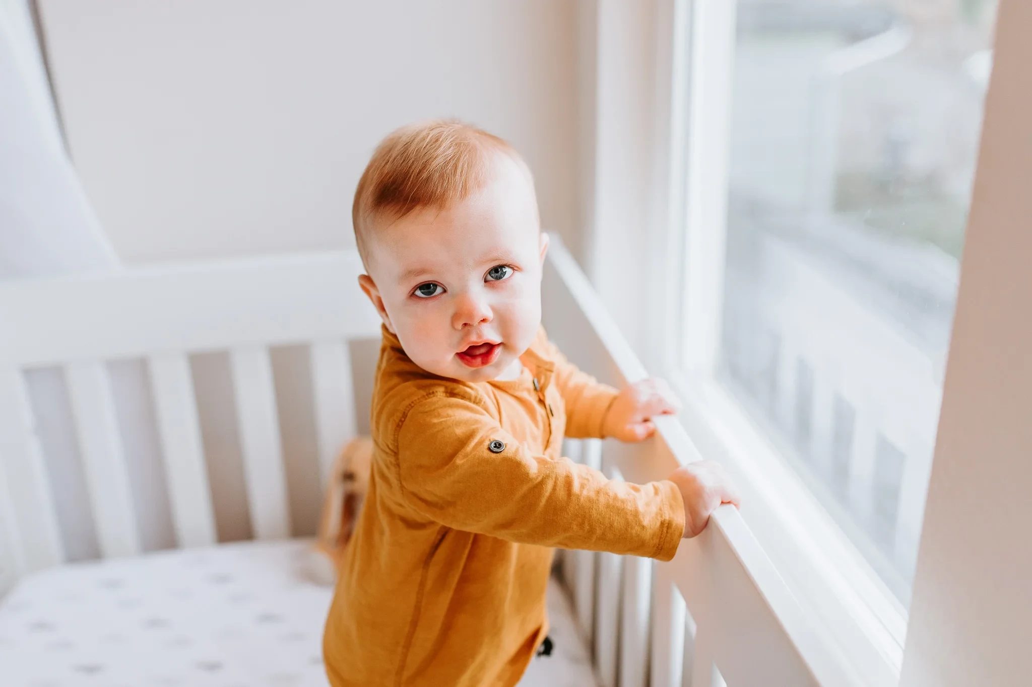 Baby in a mustard outfit stands holding a white crib rail