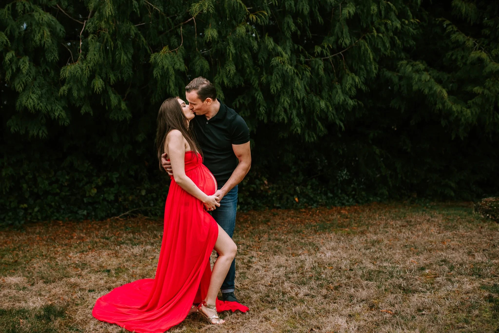 Two adults embrace in a grassy clearing, a red dress spreading across the ground
