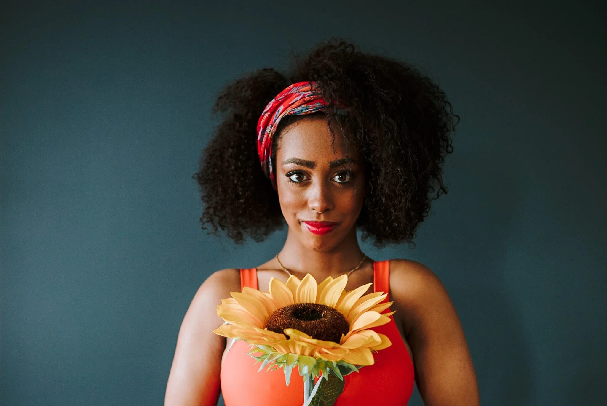 Woman in a red headband holds a sunflower against a dark green background