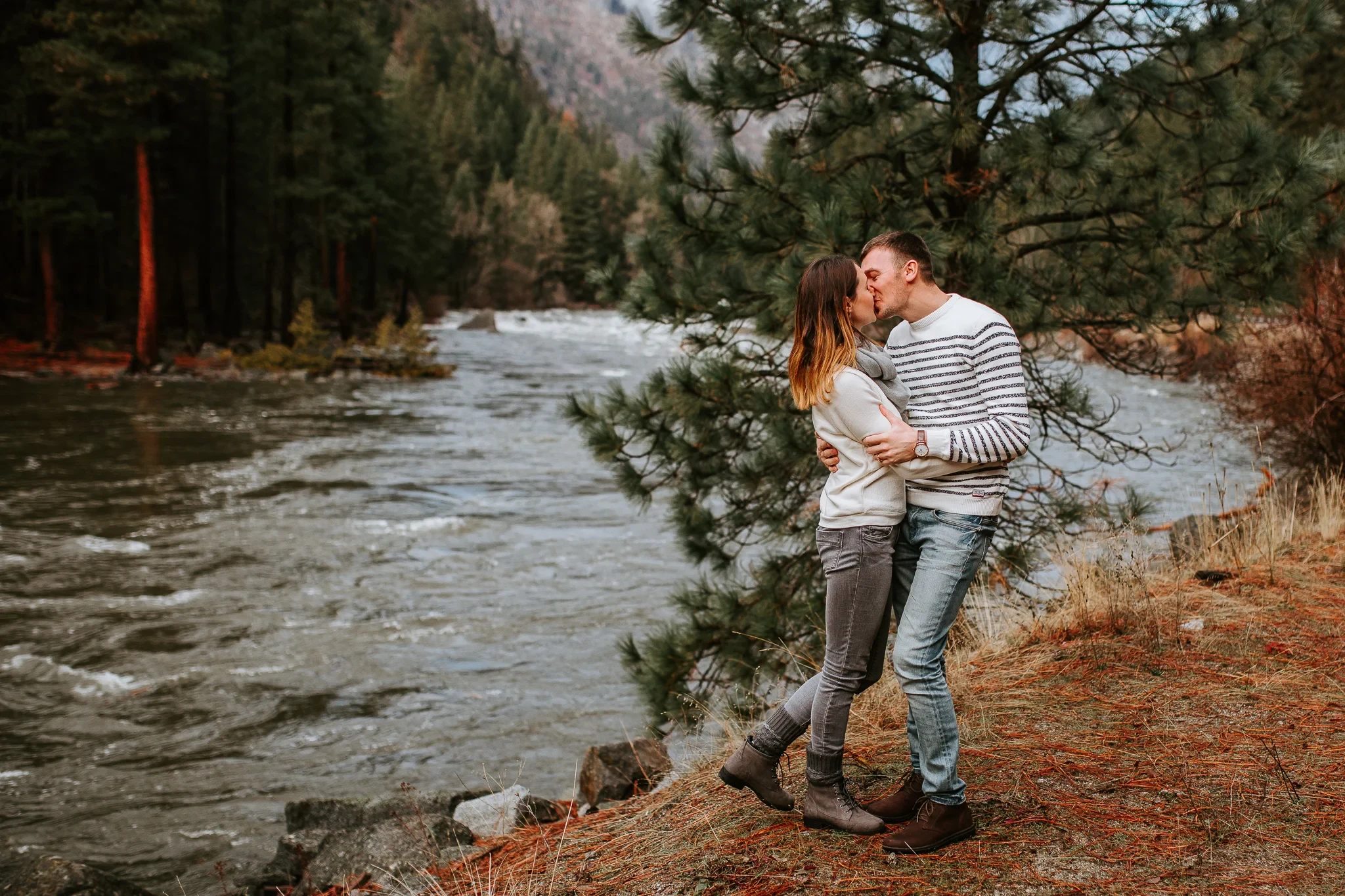 Two adults embrace beside a rushing river and evergreen woodland
