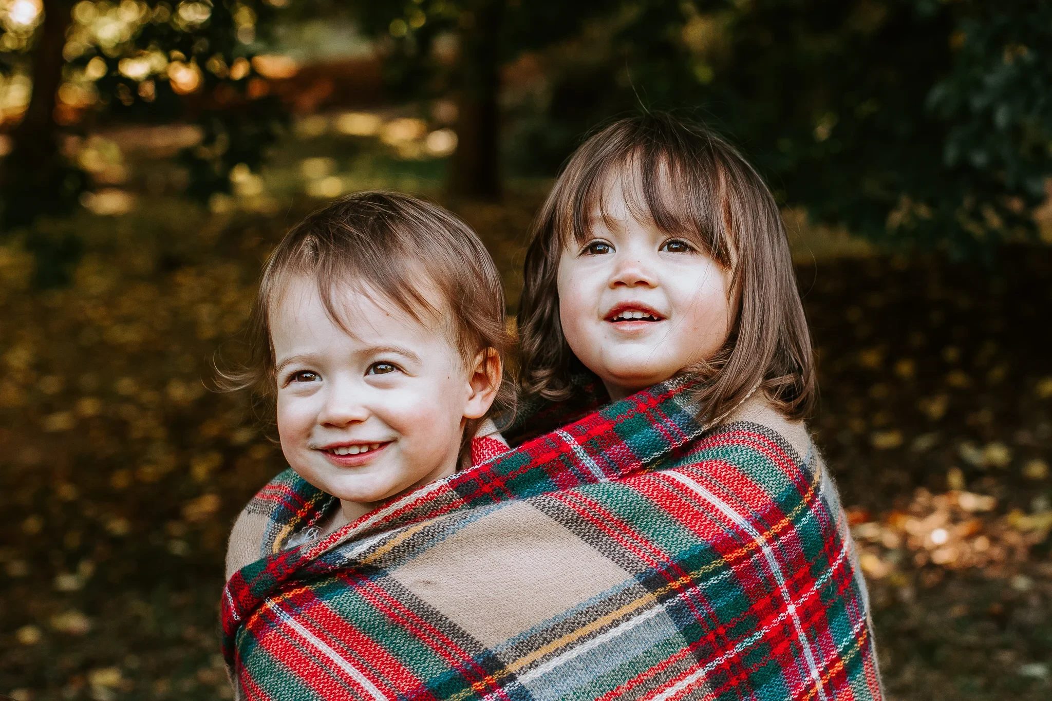 Two smiling children share a plaid blanket in the woods