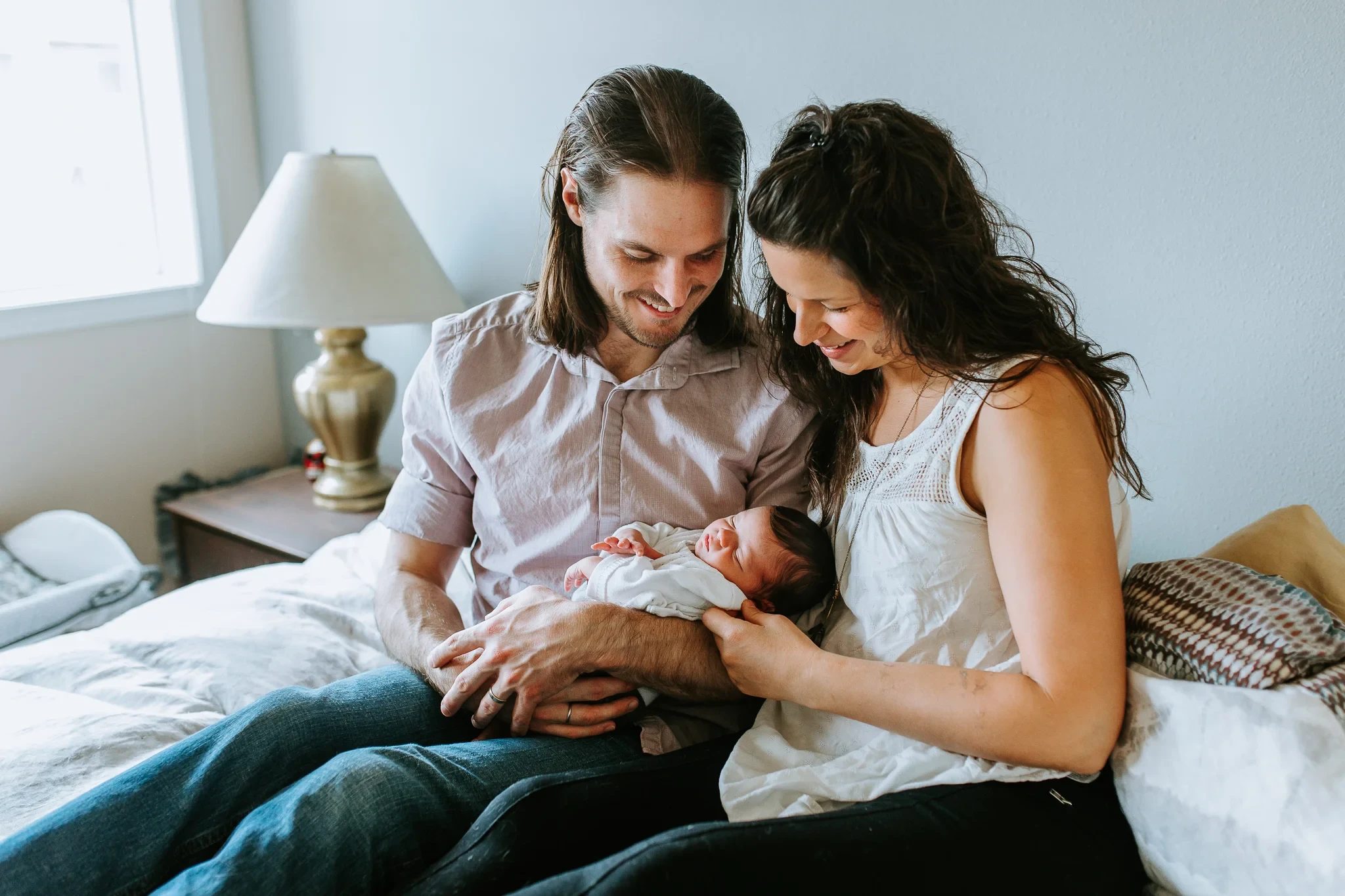 Two adults smile down at a baby while sitting on a bed