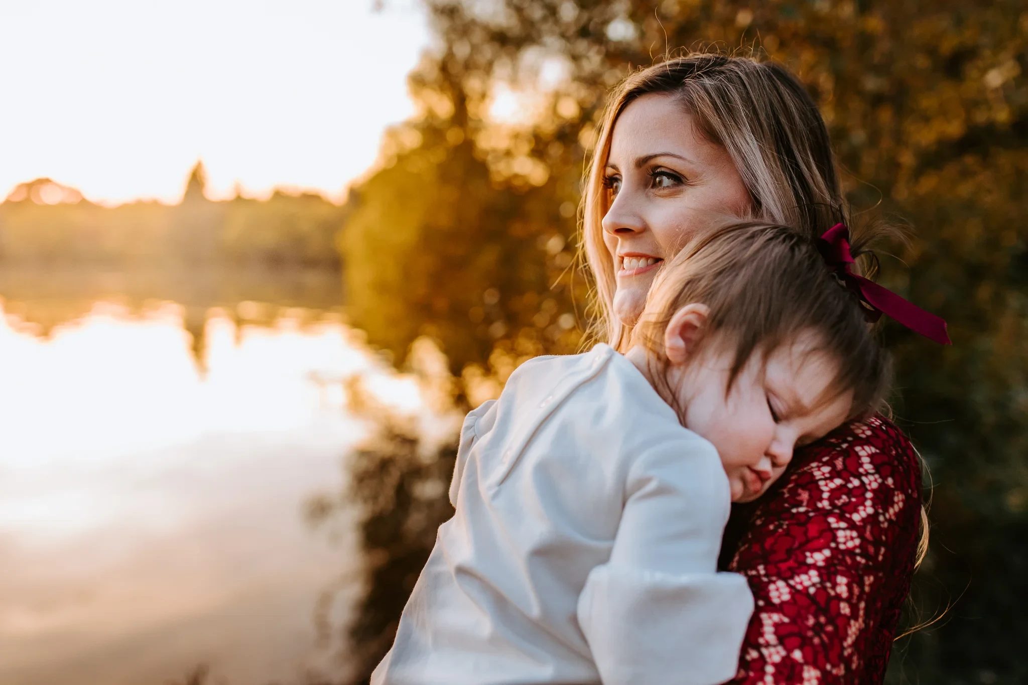 Woman cradles a resting child beside a lake at sunset