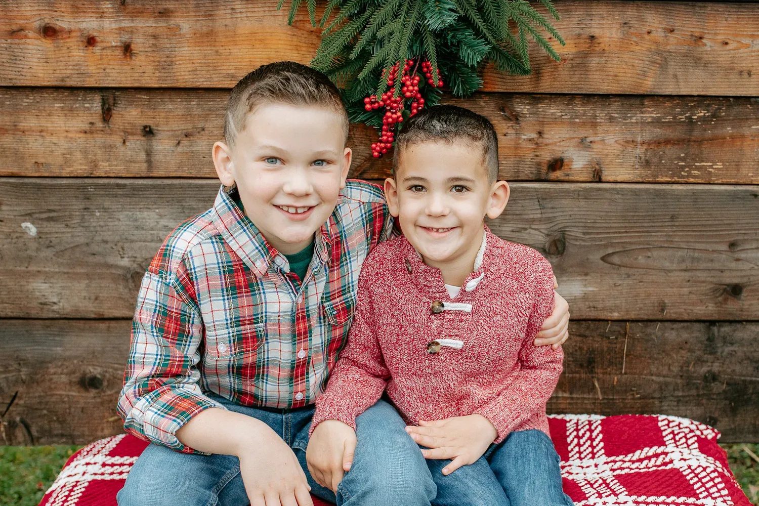 Two children sit on a plaid blanket in front of a wooden wall