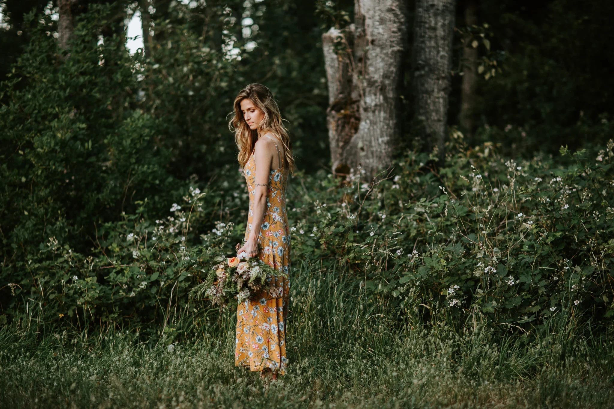 Woman in a yellow dress stands among leafy woodland shrubs