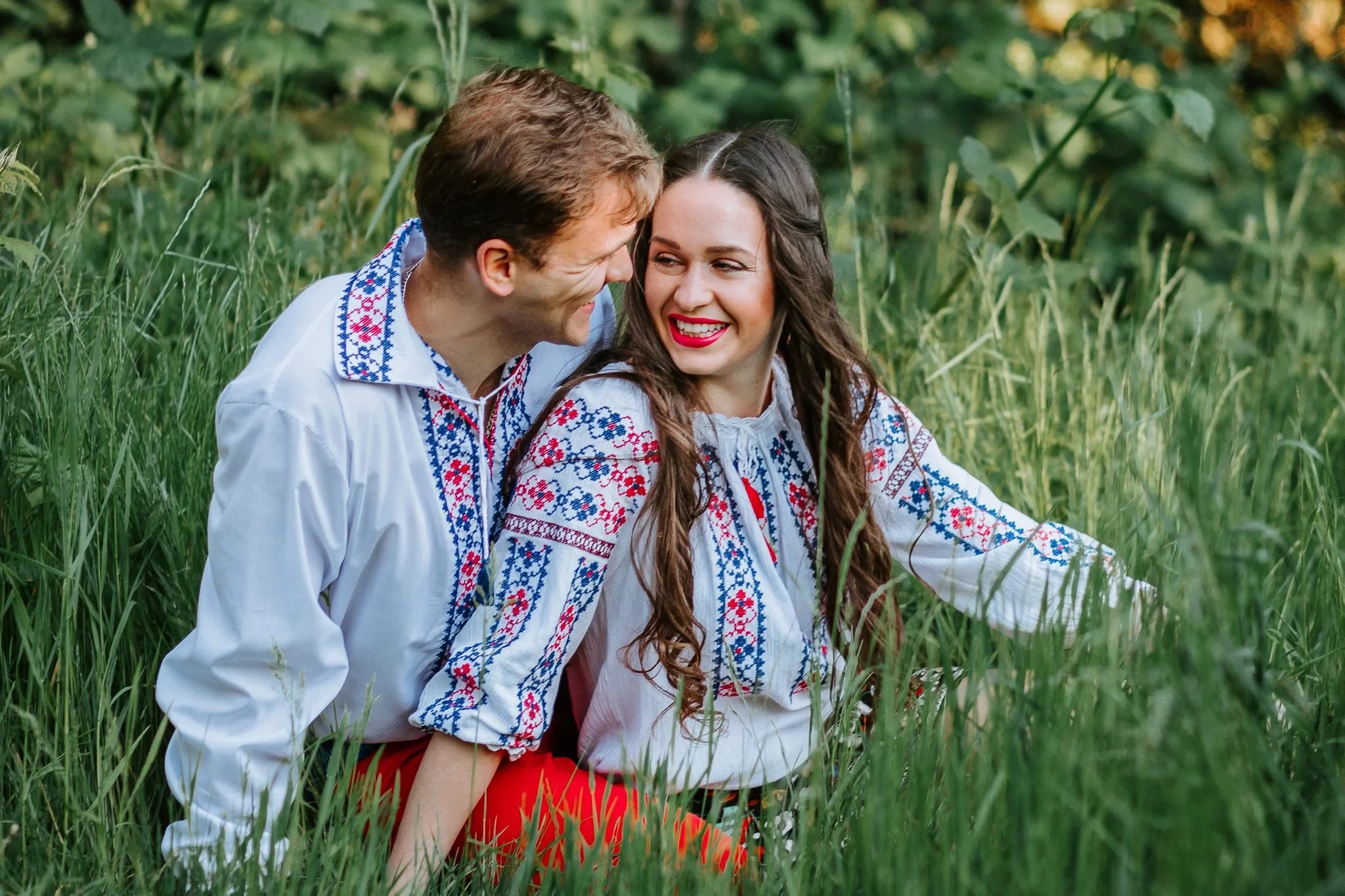 Two adults laugh together in tall grass wearing embroidered shirts