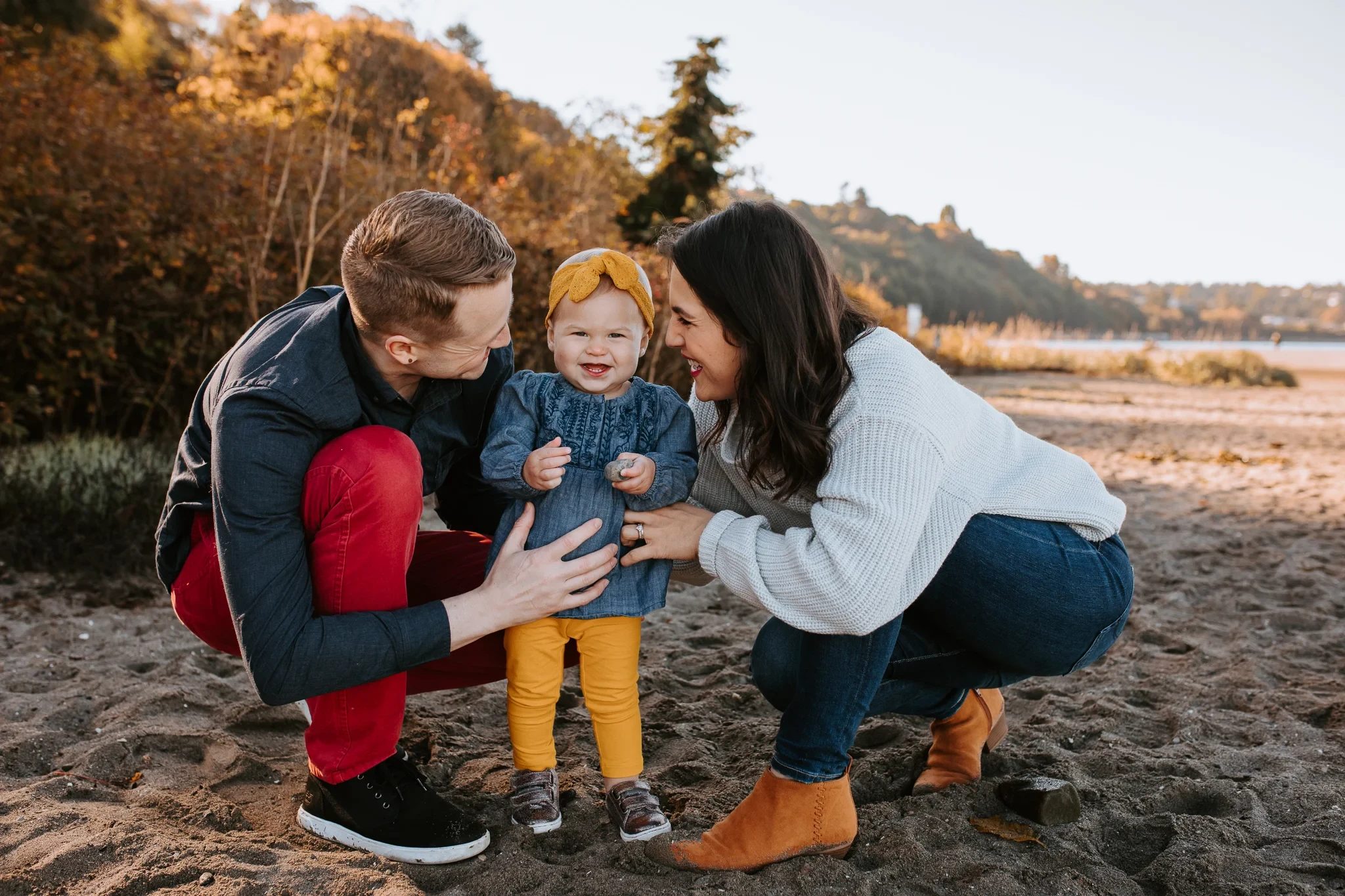 Two adults crouch beside a smiling toddler on a sandy shore