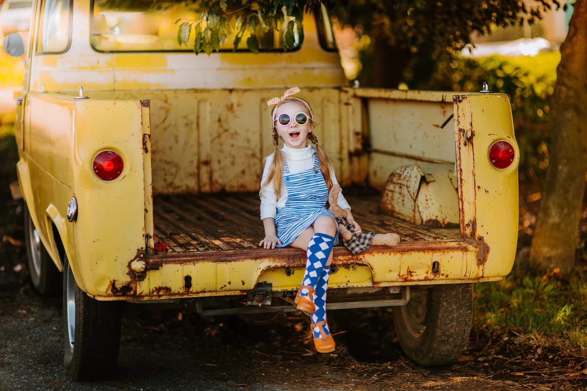Child wearing round sunglasses sits on the tailgate of a weathered yellow pickup truck