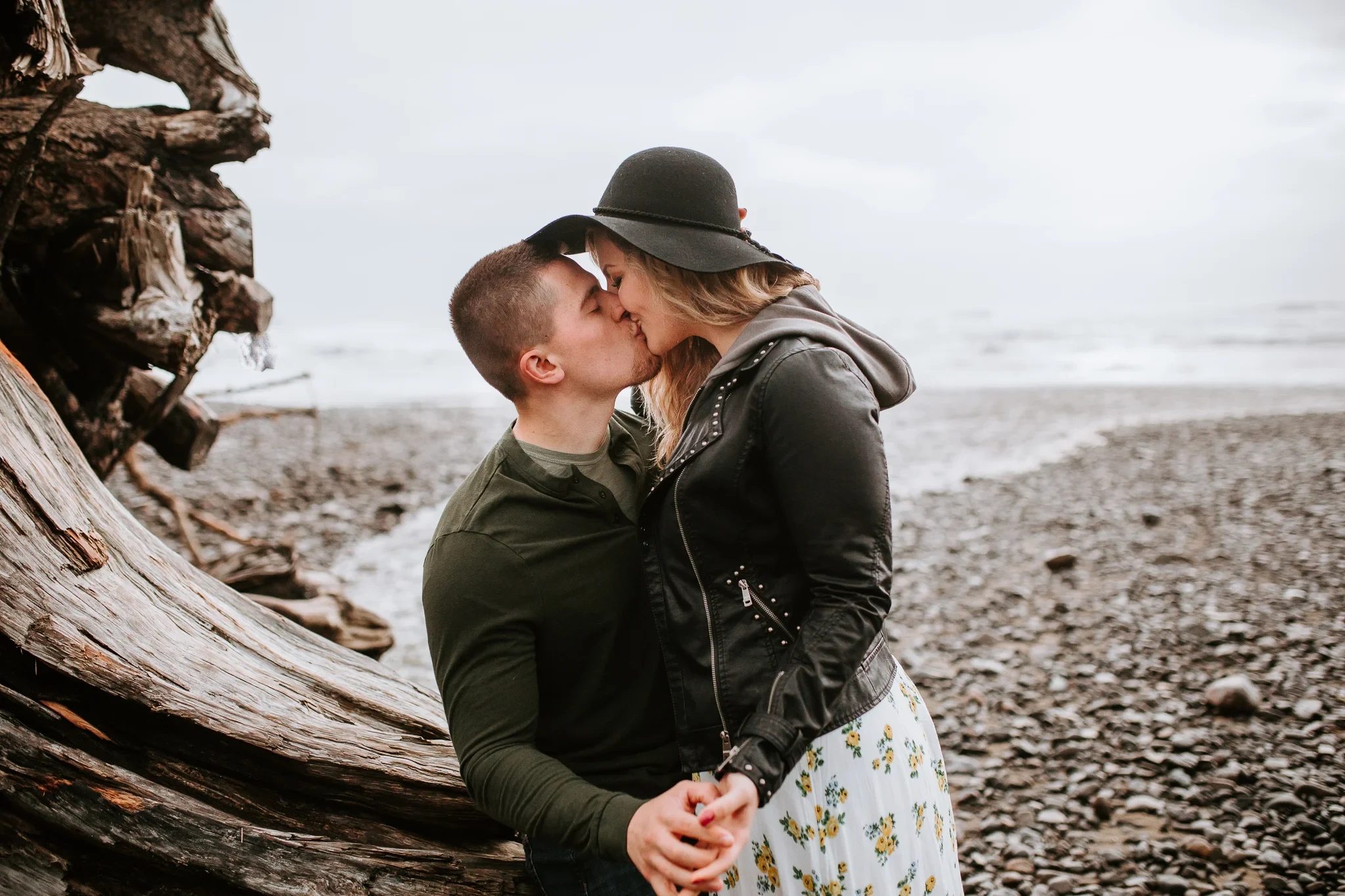 Two adults kiss beside driftwood on a pebbled beach