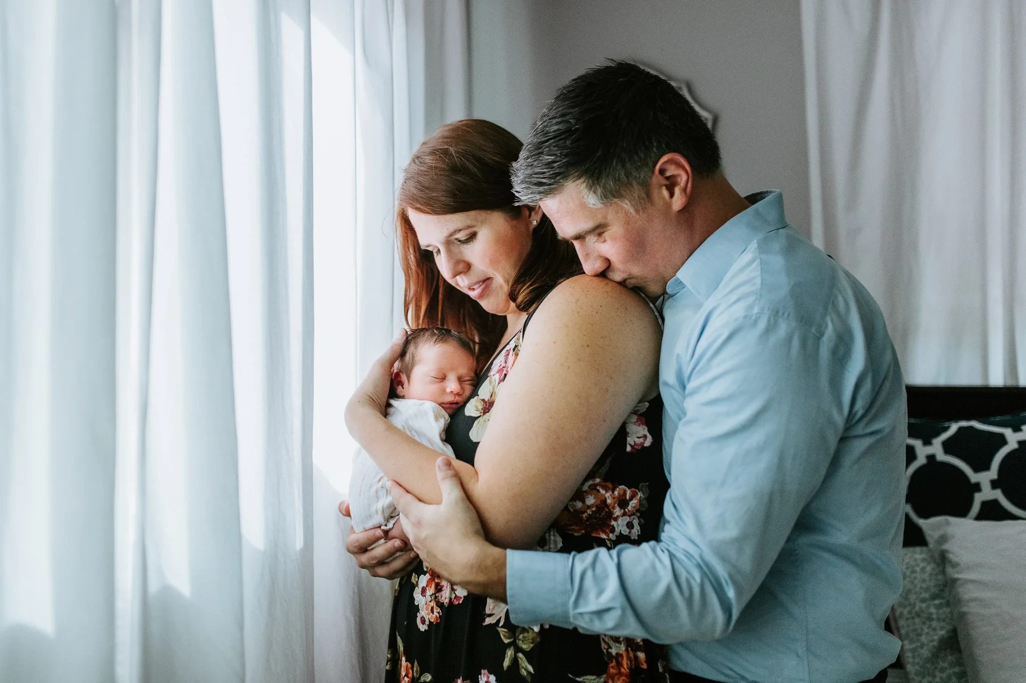 Two adults hold a baby together beside sheer curtains