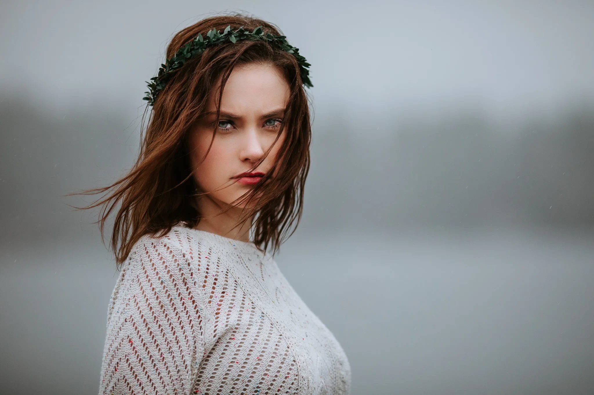 Woman wearing a leafy headband looks toward the camera beside misty water
