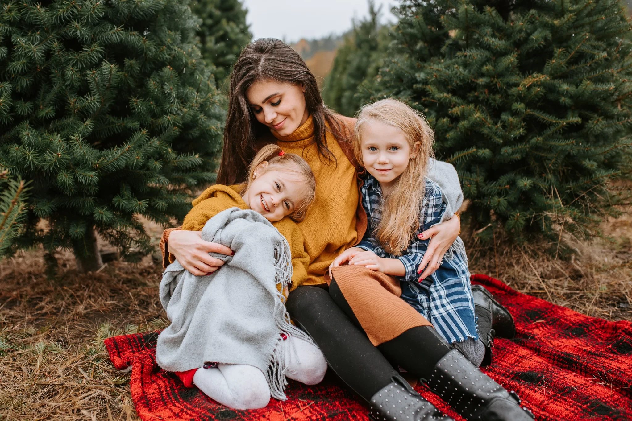 Woman and two children sit together on a red blanket among evergreen trees