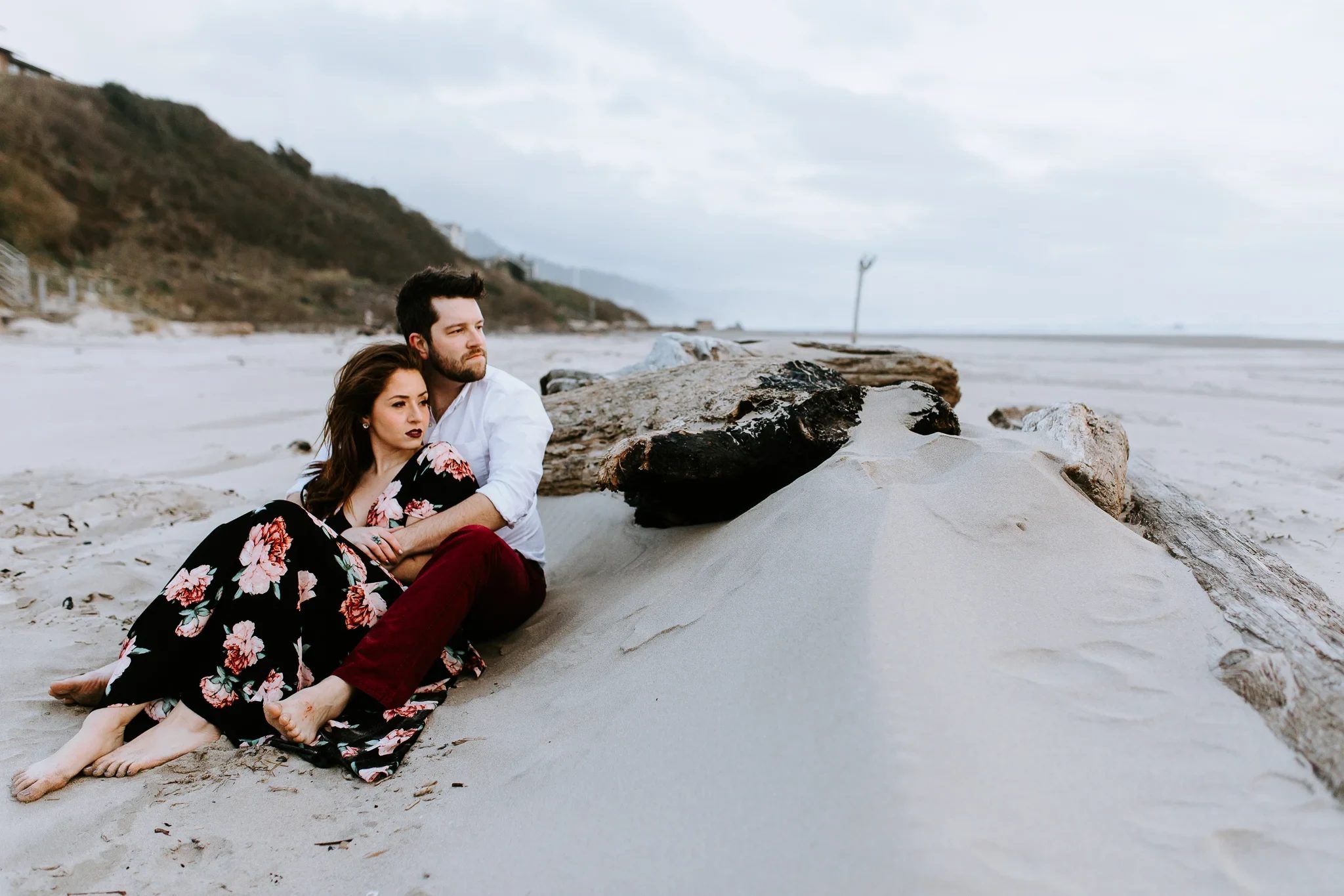 Two adults relax against a driftwood log on a sandy beach