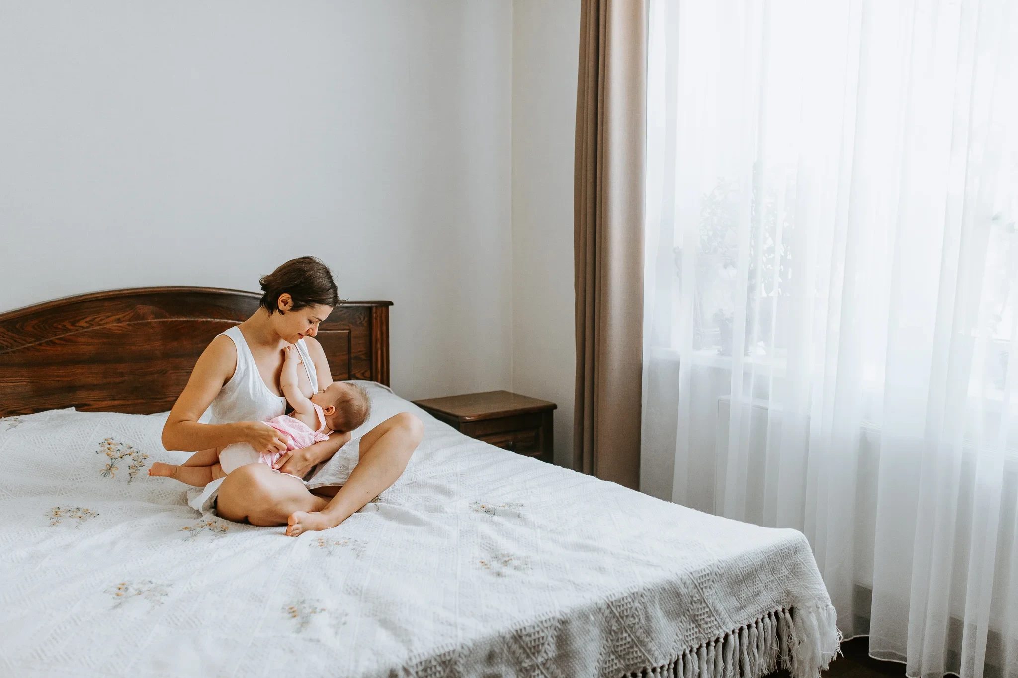 Wide bedroom view of a woman cradling a baby beside sheer curtains