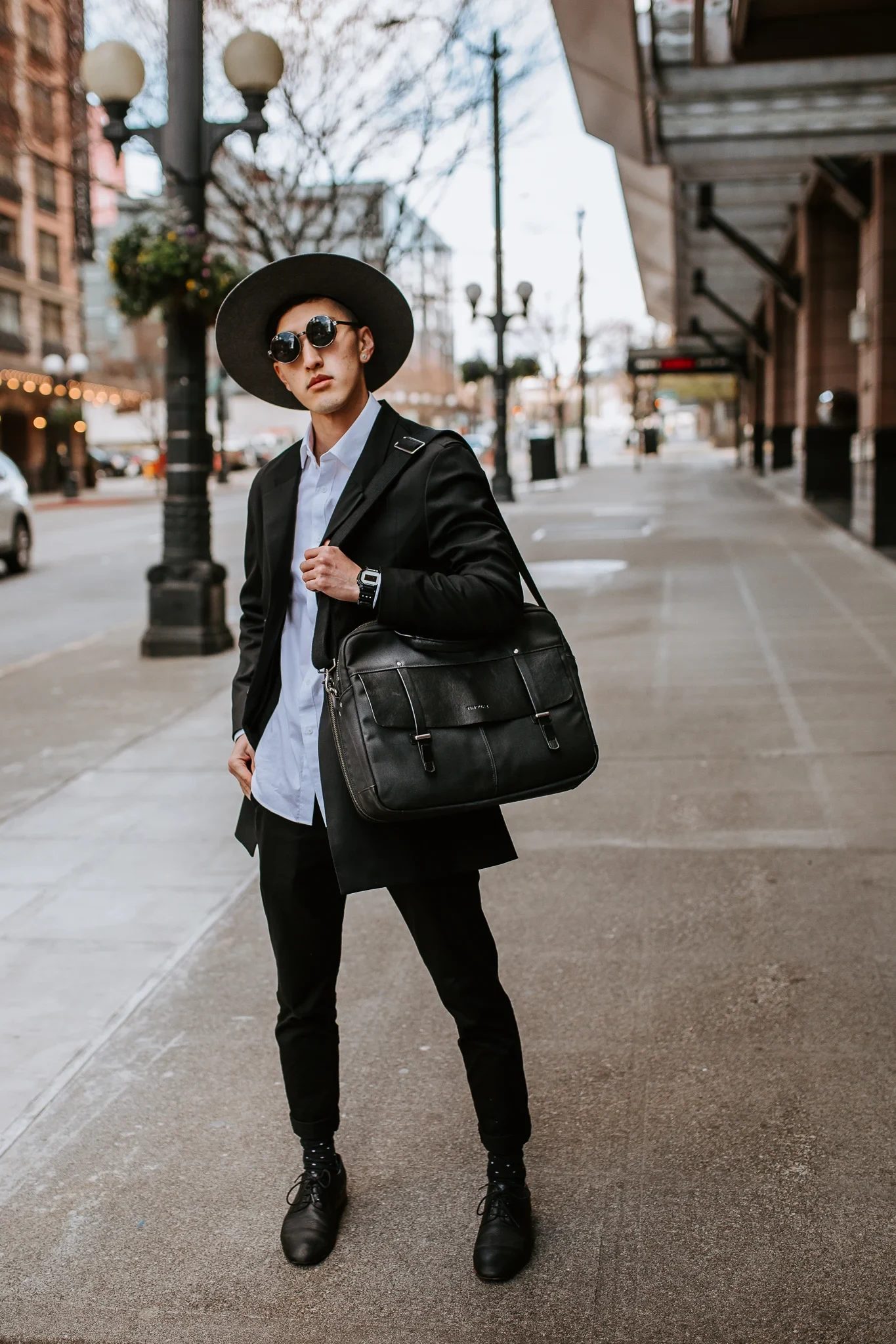 Person wearing a hat stands with a shoulder bag on a city sidewalk