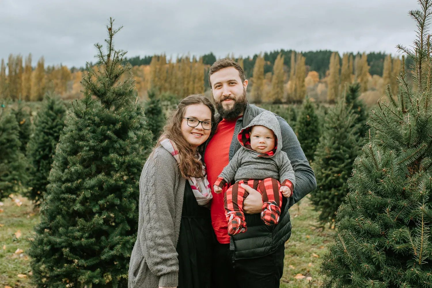 Two adults hold a bundled child in front of evergreen trees